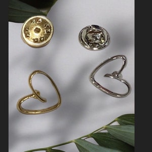 May include: Two gold and silver heart-shaped pins with a silver pin back. The pins are on a white surface with green leaves.