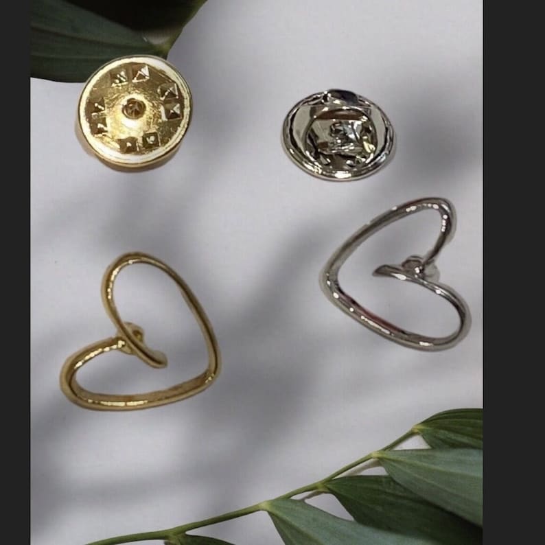 May include: Two gold and silver heart-shaped pins with a silver pin back. The pins are on a white surface with green leaves.