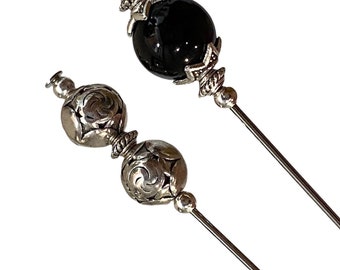 May include: Two silver and black decorative hat pins. One pin has a black glass bead with silver accents. The other pin has two silver beads with intricate designs.
