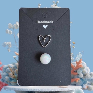 May include: A black card with the word "Handmade" and a heart symbol. The card holds a silver heart-shaped earring and a round, opalescent earring. The background is a light blue with floral and coral decorations.