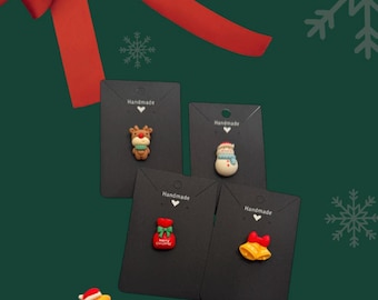 May include: A selection of Christmas-themed brooches and pins. The image shows pins in the shape of a reindeer, snowman, gift bag, bell, star, ornament, Santa Claus, and a Christmas tree. The pins are on black cards with the word "Handmade". A red bow is in the background.