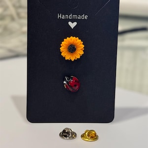 May include: A black card with the word "Handmade" and a heart symbol. The card displays a sunflower pin, a ladybug pin, and two pin backs, one silver and one gold. The sunflower pin is yellow and black, and the ladybug pin is red with black spots.