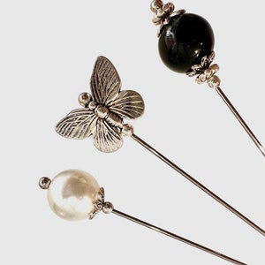 May include: Three decorative pins with different embellishments. One pin features a silver butterfly, another has a white pearl, and the third has a black gemstone.