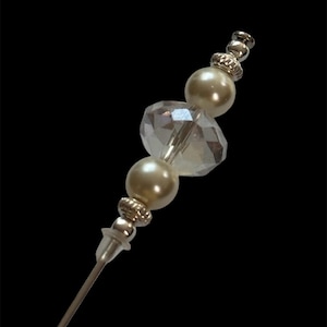 May include: A decorative silver-toned hat pin with a pearl and crystal bead design. The pin features two faux pearls, a faceted crystal bead, and decorative silver-toned accents. The pin is designed to be inserted into a hat or other fabric.