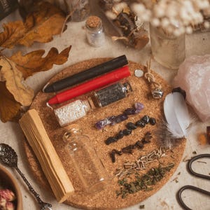 May include: A cork board with various items for a spell or ritual, including a red candle, a black candle, a small bottle of white crystals, a small bottle of black crystals, a small bottle of herbs, a feather, a piece of palo santo wood, and a small bottle of clear liquid.
