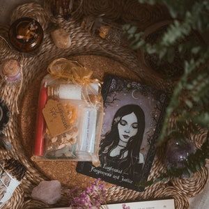 May include: A selection of spiritual items displayed on a woven mat. The items include a spell kit with red candles, a tarot card with a woman's portrait, crystals, and a burning candle in a black holder. The scene is decorated with dried herbs and flowers.
