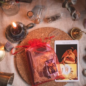 May include: A red mesh bag filled with various items, including a tarot card with the image of The Devil, a small bottle, and a crystal. The bag is sitting on a cork coaster with a handwritten note that says "The Devil Spell".