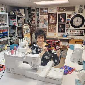 May include: A woman holding a black, red, and yellow zippered pouch with the text "CHIEFS" and team logos. The woman is in a sewing room with sewing machines and fabric.