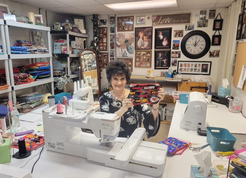 May include: A woman holding a black, red, and yellow zippered pouch with the text "CHIEFS" and team logos. The woman is in a sewing room with sewing machines and fabric.