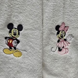 May include: White terry towels featuring embroidered images of Mickey Mouse and Minnie Mouse. Mickey is in red shorts and yellow shoes. Minnie is wearing a pink dress and bow. These towels are suitable for a bathroom or kitchen.
