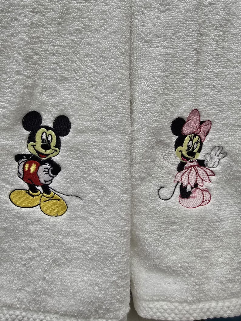 May include: White terry towels featuring embroidered images of Mickey Mouse and Minnie Mouse. Mickey is in red shorts and yellow shoes. Minnie is wearing a pink dress and bow. These towels are suitable for a bathroom or kitchen.