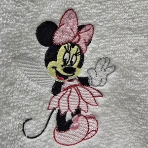 May include: Embroidered Minnie Mouse design on a white towel. The image shows Minnie Mouse in a pink dress, bow, and shoes, with a black head and ears, and a white glove. The embroidery is detailed and colourful.