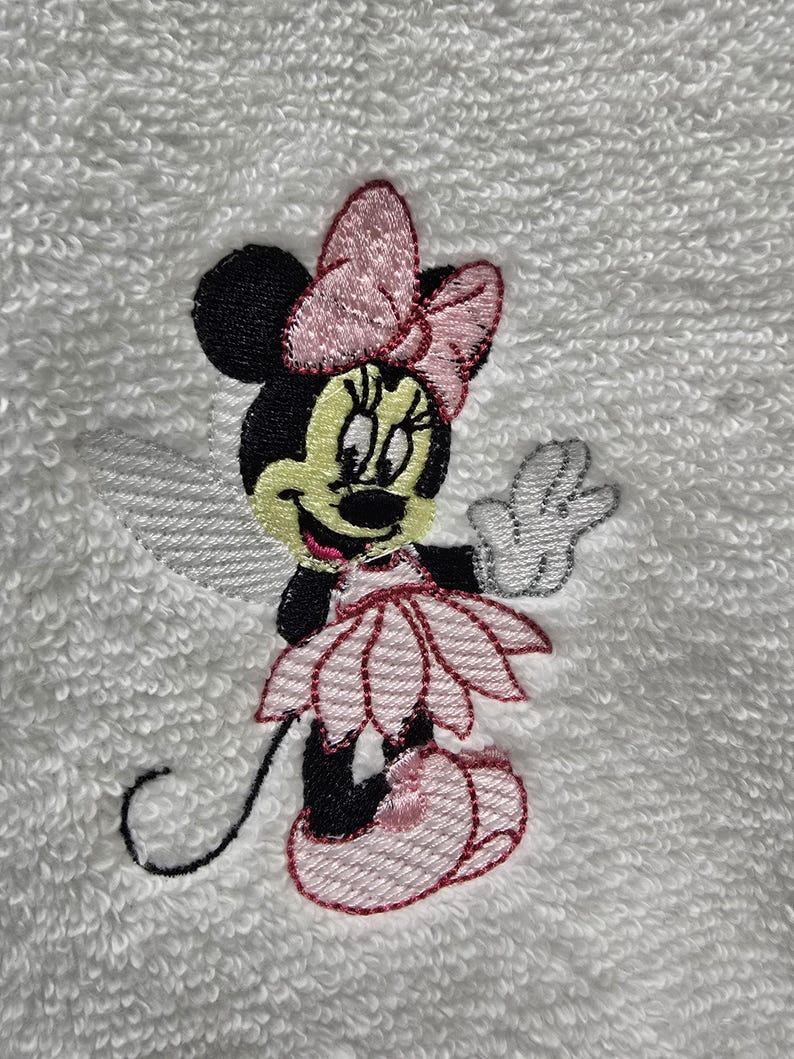 May include: Embroidered Minnie Mouse design on a white towel. The image shows Minnie Mouse in a pink dress, bow, and shoes, with a black head and ears, and a white glove. The embroidery is detailed and colourful.