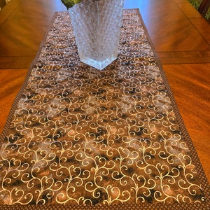 May include: A brown table runner with a gold swirl pattern. The runner has a brown border and is made of fabric.