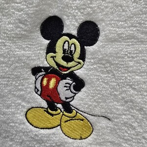 May include: Embroidered image of Mickey Mouse on a white, textured surface. Mickey is depicted in his classic outfit: black shorts, yellow shoes, white gloves, and a red and yellow vest. The image is a close-up of the character.