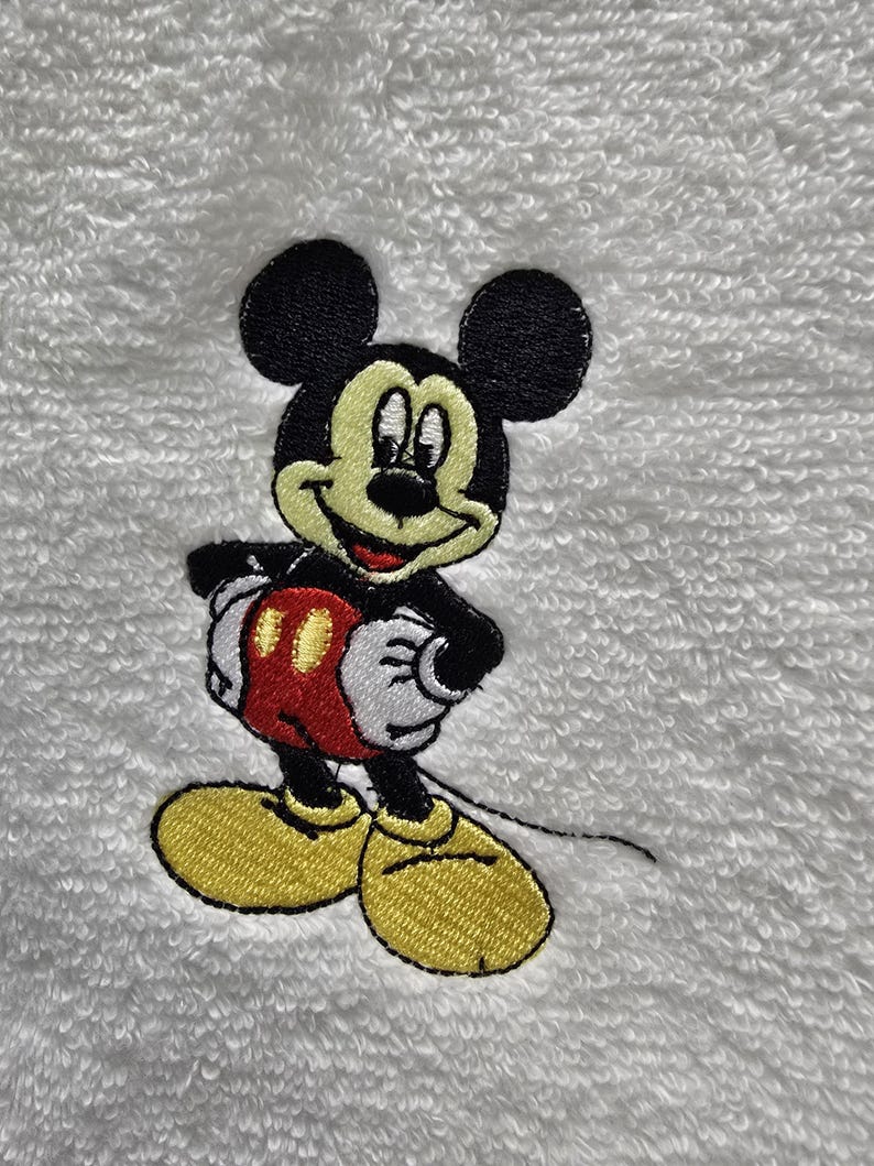 May include: Embroidered image of Mickey Mouse on a white, textured surface. Mickey is depicted in his classic outfit: black shorts, yellow shoes, white gloves, and a red and yellow vest. The image is a close-up of the character.