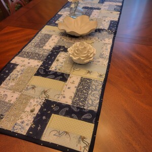 May include: A blue and white patchwork table runner with a dark blue border. The runner features various patterns, including floral, paisley, and bicycle motifs. Decorative white ceramic bowls and a vase with flowers sit on top of the runner.