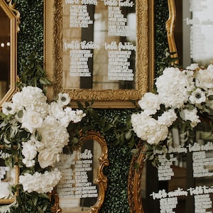 May include: A wall covered in green ivy with gold framed mirrors displaying white handwritten seating charts for a wedding reception. The mirrors are decorated with white flowers.