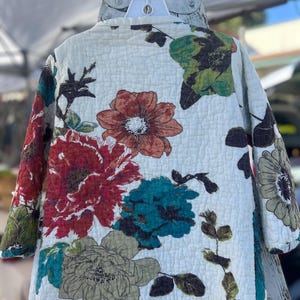 May include: A quilted jacket with a white background and large floral patterns in shades of red, teal, brown, and green. The jacket is hanging on a white hanger, showcasing its design and texture. The floral print adds a touch of nature-inspired style.