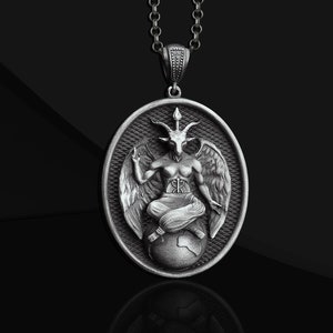 Può includere: Una collana con un ciondolo d'argento che raffigura una figura cornuta seduta su un globo. La figura ha le ali e indossa un perizoma. Il ciondolo è circolare e ha una superficie testurizzata.
