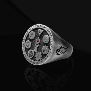Puede incluir: Un anillo plateado con una cara circular que presenta varios símbolos y una pequeña gema roja. El anillo tiene texto grabado alrededor del borde y un diseño estilizado en el lateral. El anillo se muestra sobre una superficie negra.