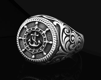 Puede incluir: Un anillo de plata con un ancla y un diseño de rueda de barco. El anillo tiene un diseño detallado y ornamentado.