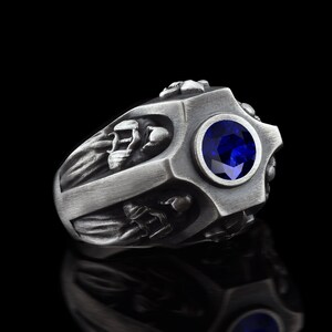 Könnte beinhalten: Ein silberner Ring mit einem blauen Edelstein und filigranen Details. Der Ring hat ein Kreuzdesign mit Figuren auf jeder Seite.