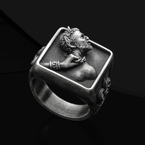 May include: A silver ring with a square design featuring a detailed sculpture of a man with horns and a chain around his neck.