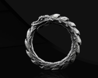 May include: A silver ring shaped like a dragon biting its tail, creating a circle. The ring is on a black background.