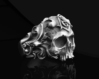 May include: A silver skull ring featuring intricate details and a Masonic symbol on the top. The ring is set against a black background.
