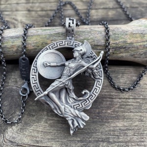 May include: A silver necklace with a pendant featuring a Greek goddess holding a spear and shield. The pendant is surrounded by a circular design with a Greek key pattern.