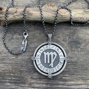 May include: Silver necklace with a round pendant featuring the Virgo zodiac sign surrounded by the other zodiac signs. The pendant has a textured, aged look and is attached to a silver chain.