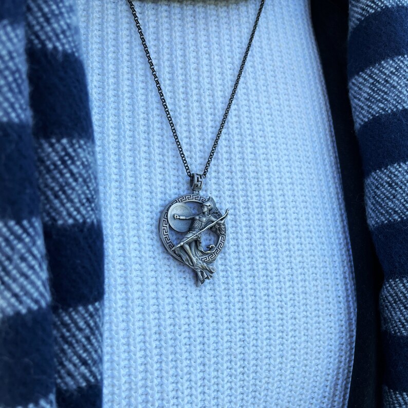 May include: A silver necklace with a pendant featuring a detailed depiction of a woman holding a spear and shield, surrounded by a circular Greek key design.
