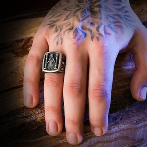 May include: A silver ring with a Masonic symbol, featuring a pyramid with an all-seeing eye, two pillars, and other intricate details.