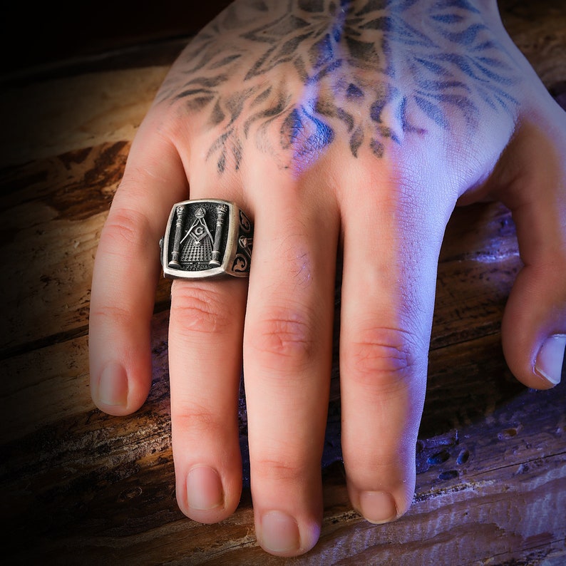May include: A silver ring with a Masonic symbol, featuring a pyramid with an all-seeing eye, two pillars, and other intricate details.