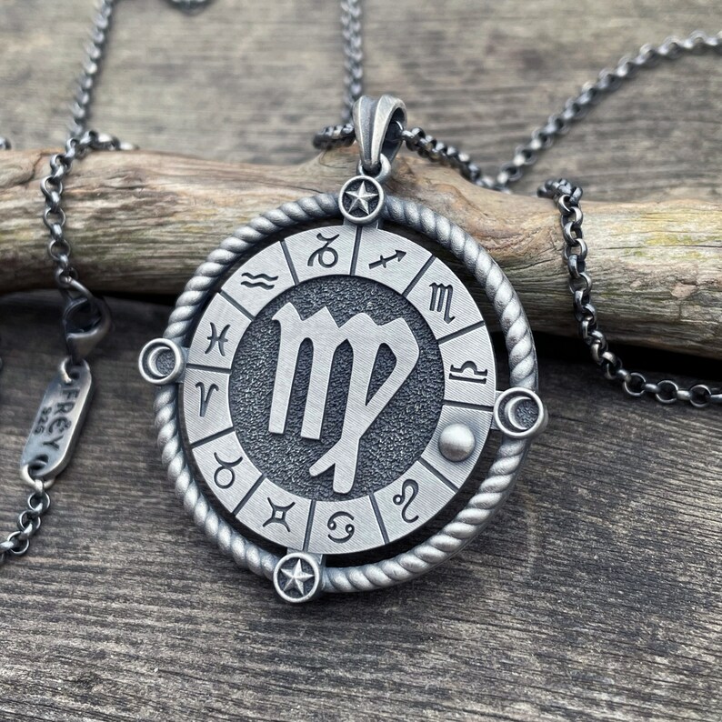 May include: A silver necklace with a round pendant featuring the Virgo zodiac sign surrounded by a circle of all the zodiac signs. The pendant is on a silver chain.