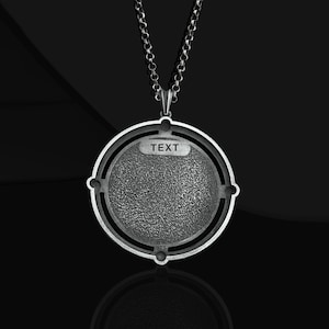 May include: A silver necklace with a round pendant featuring a textured surface and the word "TEXT" inscribed on it. The pendant is marked with "925" on the back.