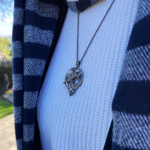 May include: A silver necklace with a pendant featuring a winged figure holding a disc. The pendant is surrounded by a circular design with a Greek key pattern.
