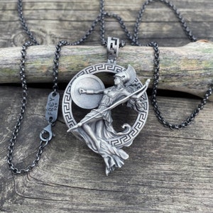 May include: Silver necklace with a pendant featuring a stylized depiction of a female warrior holding a spear and shield. The pendant is surrounded by a circular Greek key design. The necklace is made by Frey 925.