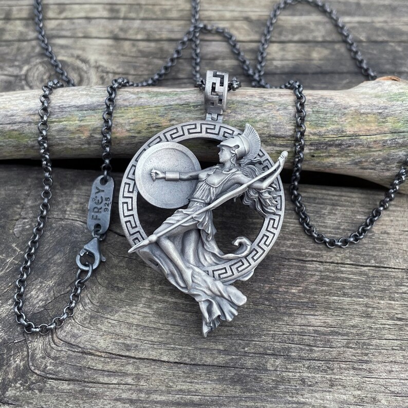 May include: Silver necklace with a pendant featuring a stylized depiction of a female warrior holding a spear and shield. The pendant is surrounded by a circular Greek key design. The necklace is made by Frey 925.