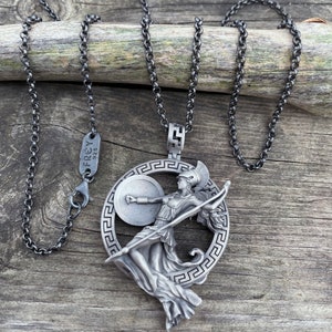 May include: A silver necklace with a pendant featuring a detailed depiction of a Greek goddess holding a spear and shield. The pendant is surrounded by a circular design with a Greek key pattern. The necklace is on a wooden surface.