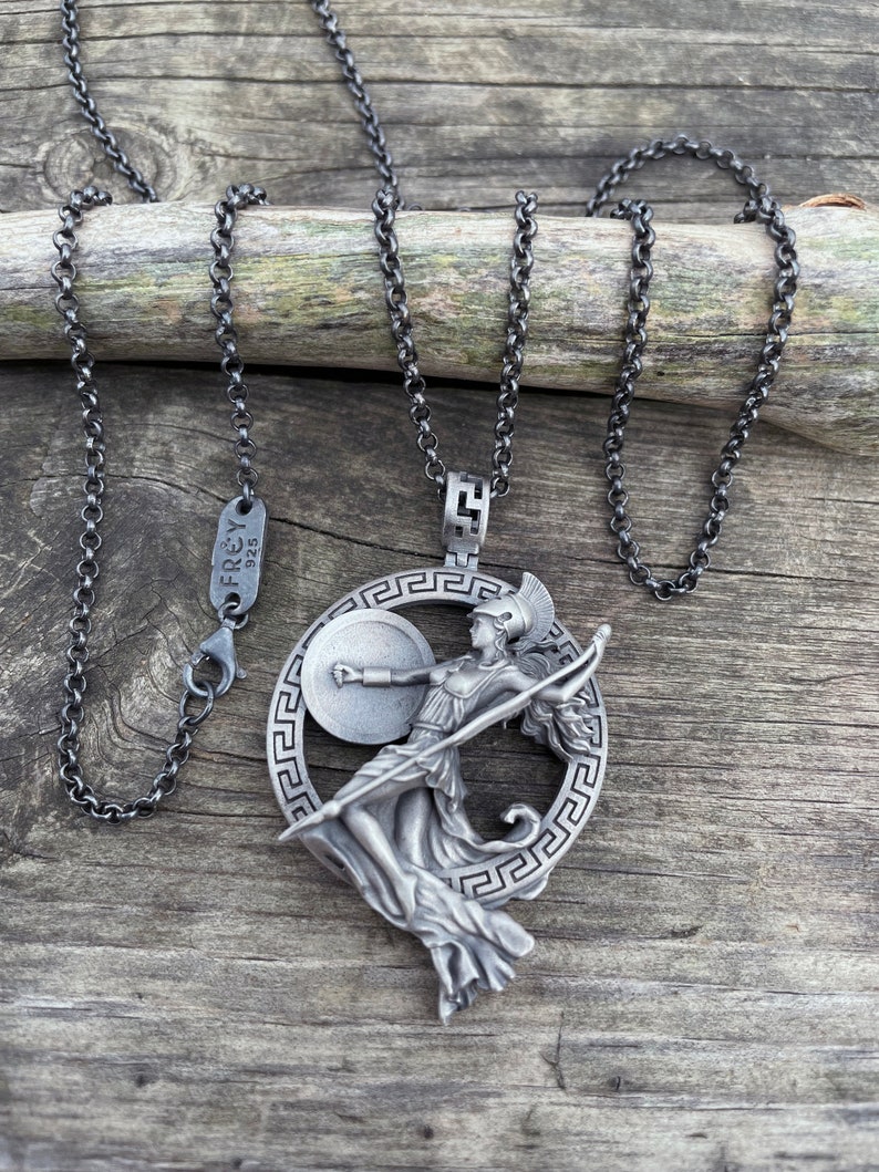May include: A silver necklace with a pendant featuring a detailed depiction of a Greek goddess holding a spear and shield. The pendant is surrounded by a circular design with a Greek key pattern. The necklace is on a wooden surface.