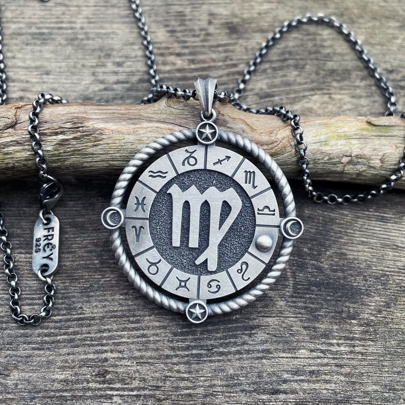 May include: A silver necklace with a round pendant featuring the Virgo zodiac sign surrounded by the other zodiac signs. The pendant has a rope-like border and a star at the top. The necklace is on a wooden surface.