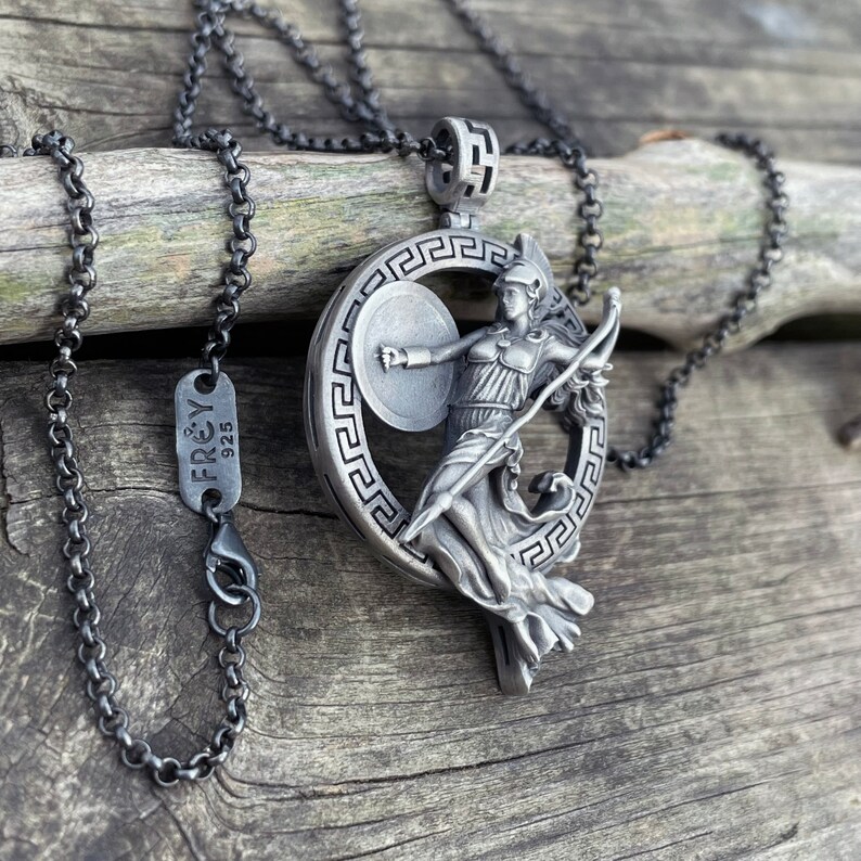 May include: A silver necklace with a pendant featuring a detailed sculpture of a woman holding a spear and shield. The pendant is surrounded by a circular design with a Greek key pattern. The necklace is on a black chain and has a tag that reads "FREY 925".