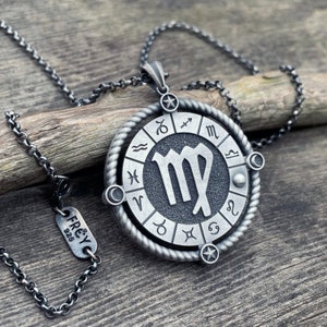 May include: A silver necklace with a round pendant featuring the astrological sign Virgo surrounded by a circle of all the zodiac signs. The pendant has a rope-like design around the edge. The necklace is on a wooden surface.
