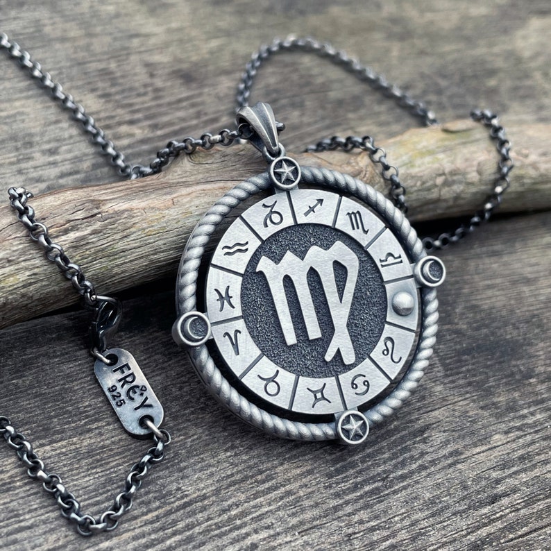 May include: A silver necklace with a round pendant featuring the astrological sign Virgo surrounded by a circle of all the zodiac signs. The pendant has a rope-like design around the edge. The necklace is on a wooden surface.