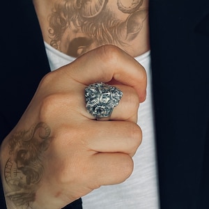 May include: A silver ring with a detailed design of a face with horns. The ring is worn on the index finger of a person's hand.