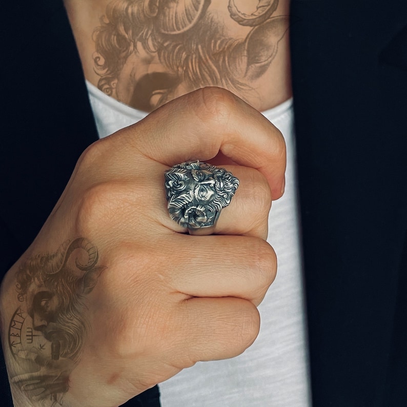 May include: A silver ring with a detailed design of a face with horns. The ring is worn on the index finger of a person's hand.