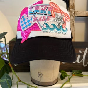 Può includere: Cappellino da camionista bianco e nero con la scritta "LAKE BABE" in rosa e blu. Il cappello presenta un cuore di paillettes rosa, una stella argentata e onde ricamate. Il cappello è esposto su una testa di manichino.