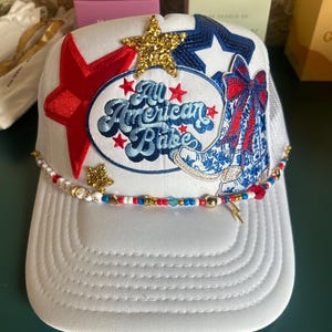 May include: A white trucker hat featuring a central oval patch with the text All American Babe, a red star, a gold glitter star, a blue sequin star, and an embroidered cowboy boot with a red and blue bow. A beaded trim with charms lines the brim.